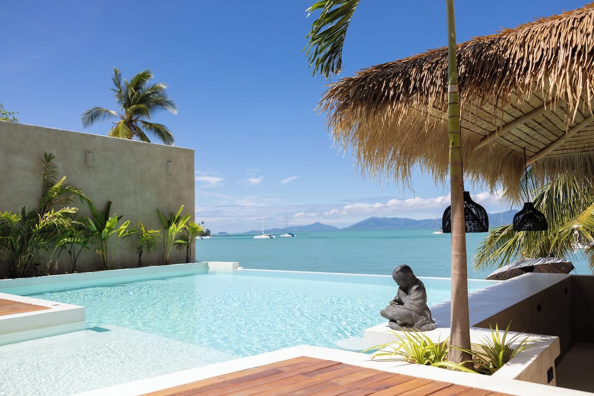 A pool area with a view of the sea, a thatched umbrella, and a stone sculpture.