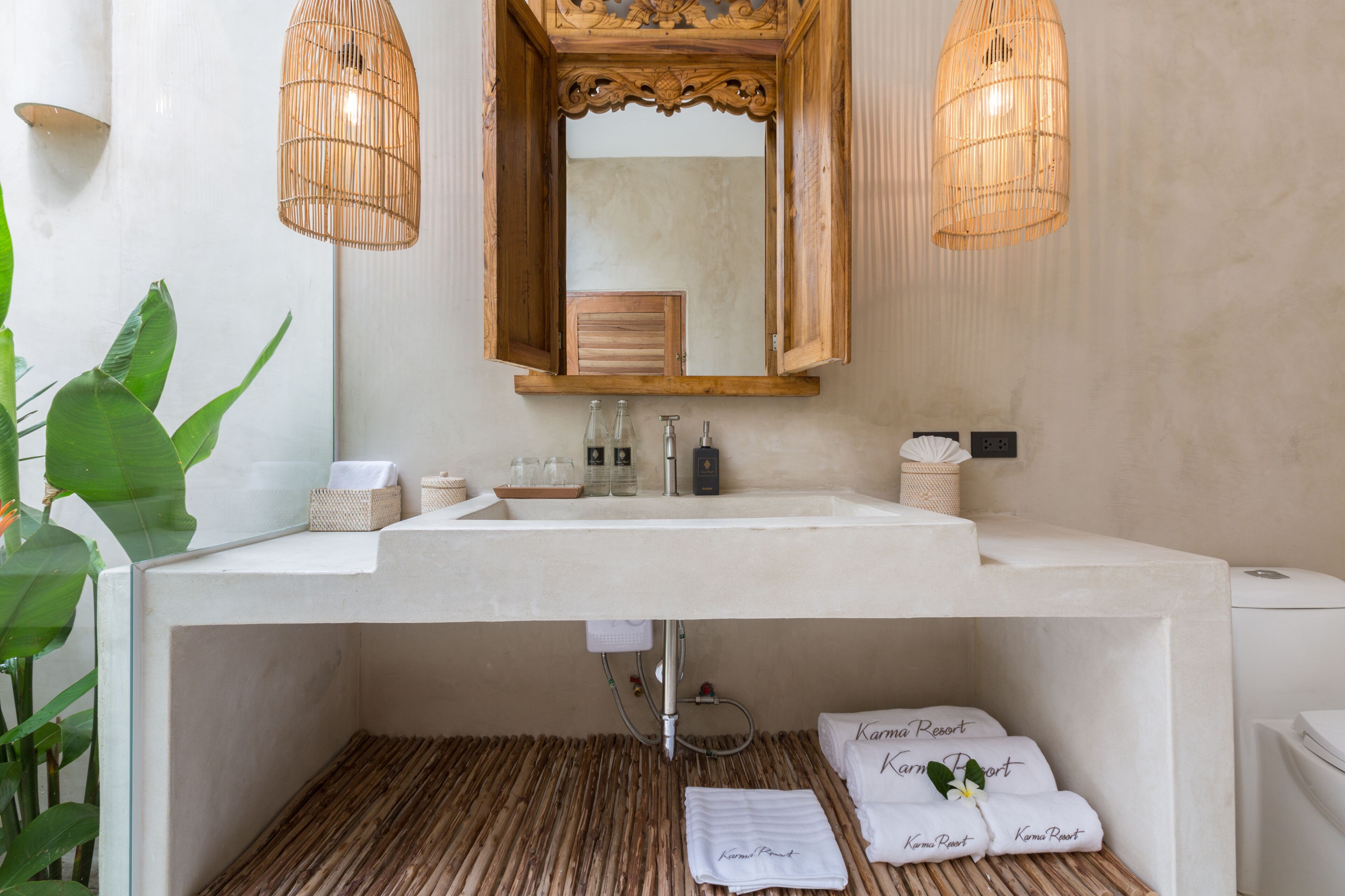 A bathroom with a white sink, wooden vanity, and a mirror above it. There are two wicker lamps, a plant, and towels with "Karma Resort" written on them.