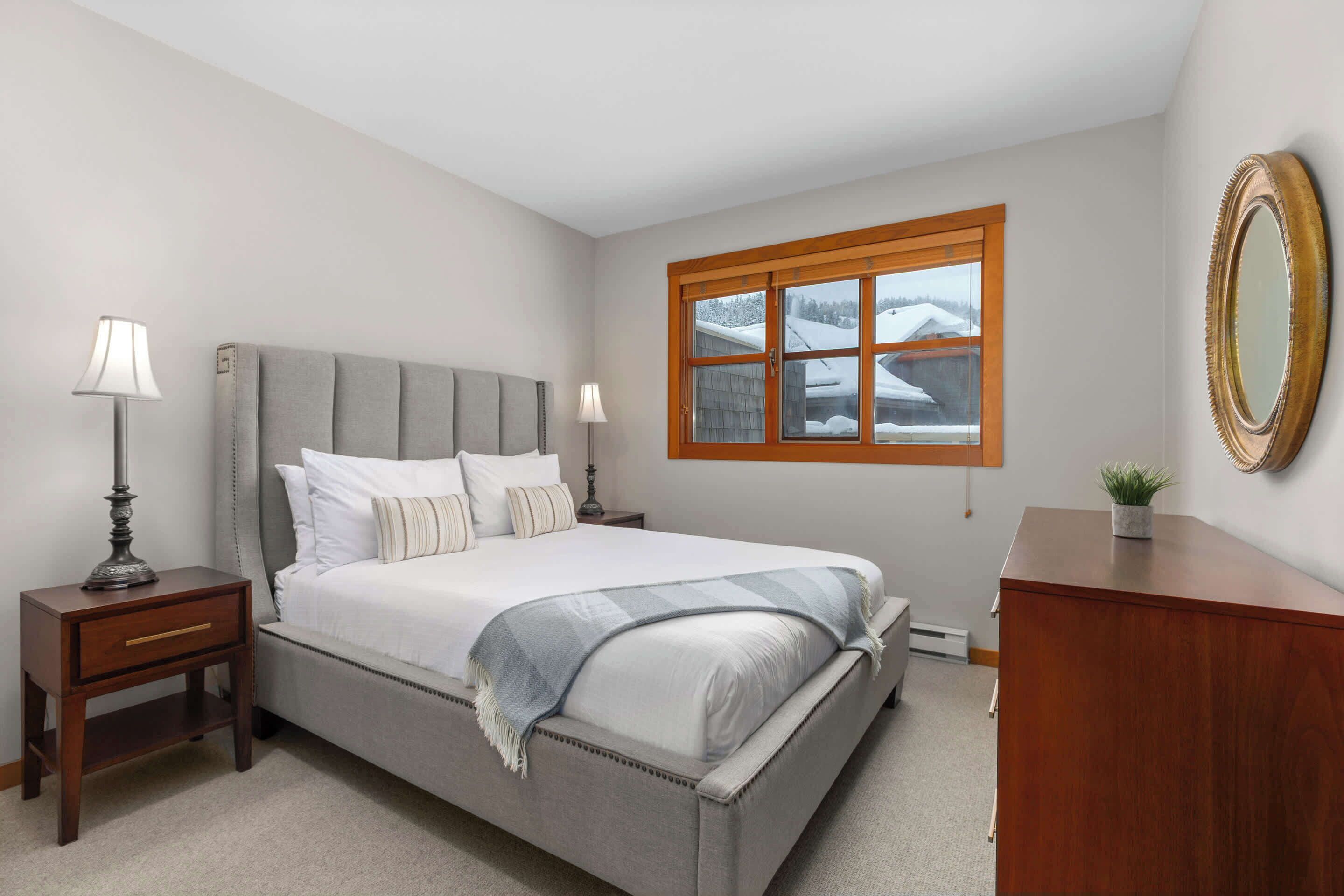 Une chambre à coucher comprenant un lit, deux tables de chevet, une commode, une coiffeuse, une fenêtre donnant sur un paysage enneigé et un miroir accroché au mur.