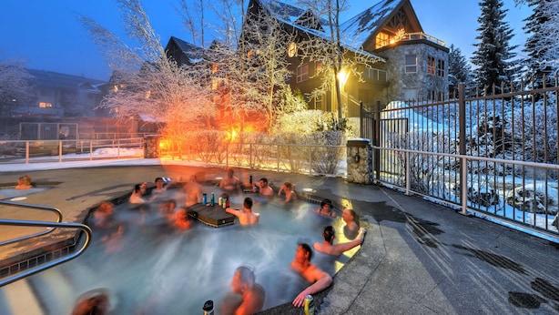 A hot tub area with people relaxing, surrounded by snow-covered trees and a stone building with a wooden roof. - Aspens #332 2-Bedroom Ski in Ski out, sleeps 6, Free Wifi (Whistler)