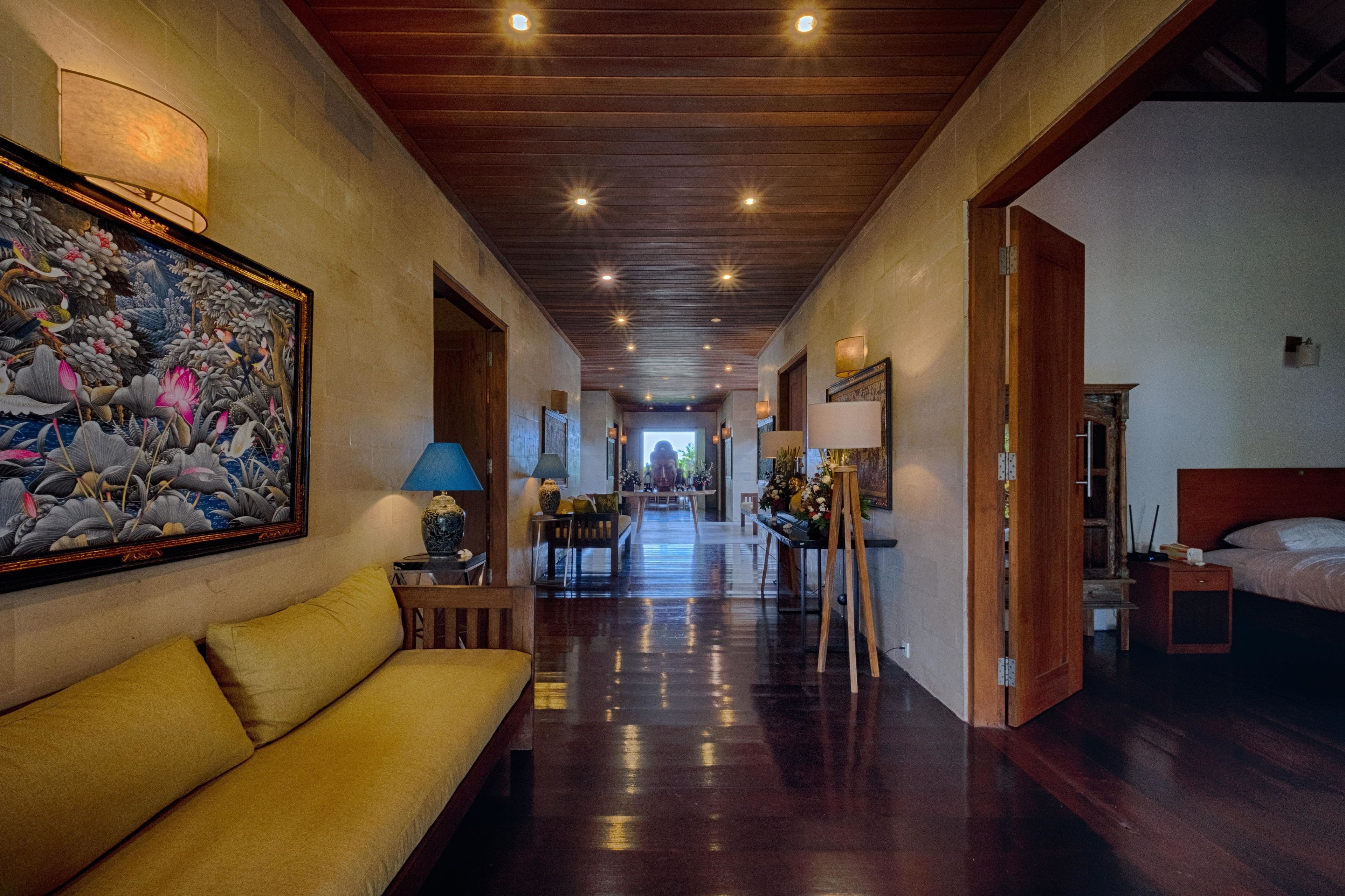 A hotel hallway with a wooden ceiling, a large painting on the left, a yellow sofa, and a bed on the right.