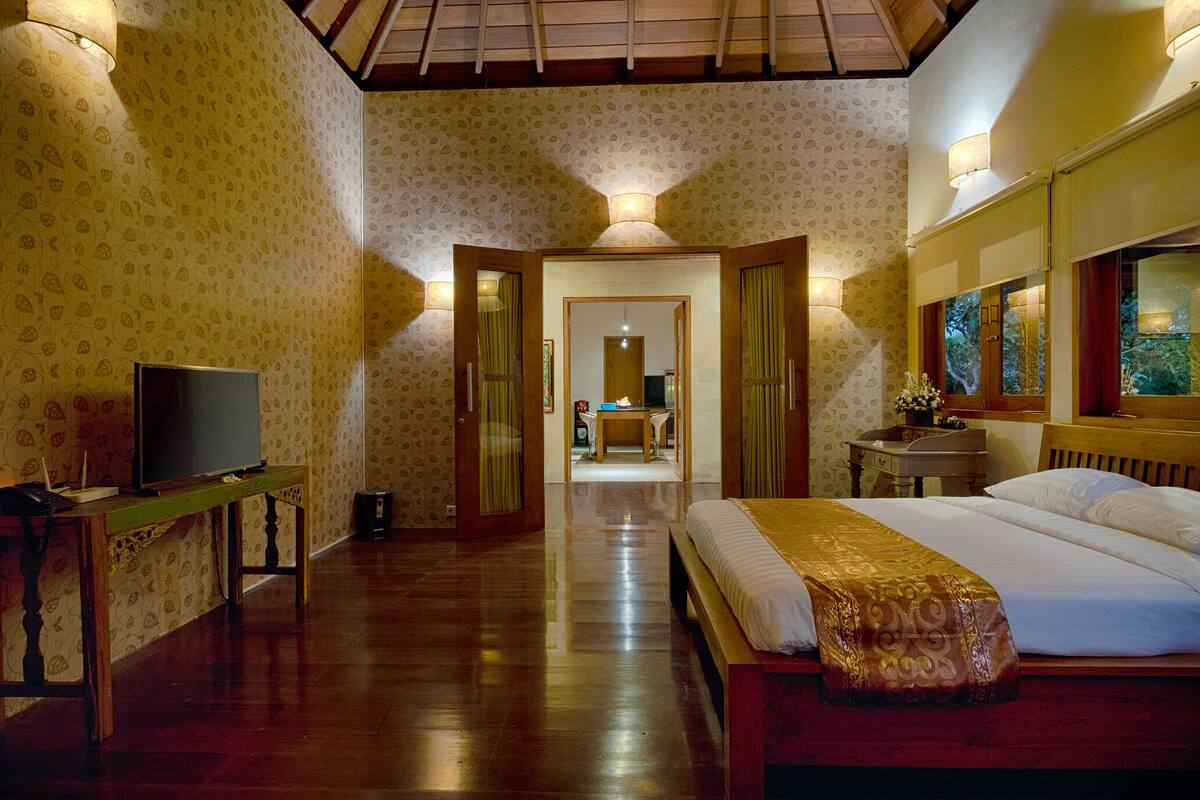Een hotelkamer met houten vloer, een bed met een gedessineerd bedovertrek, een televisie op een houten tafel en een gang die naar een andere kamer leidt.