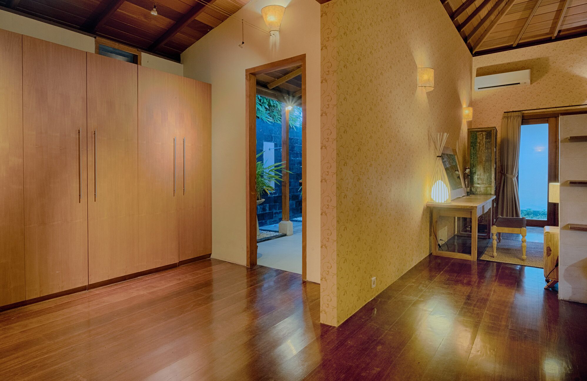Habitación amplia con piso de madera, un armario grande, una mesita con lámpara y vista a un jardín a través de un vano.