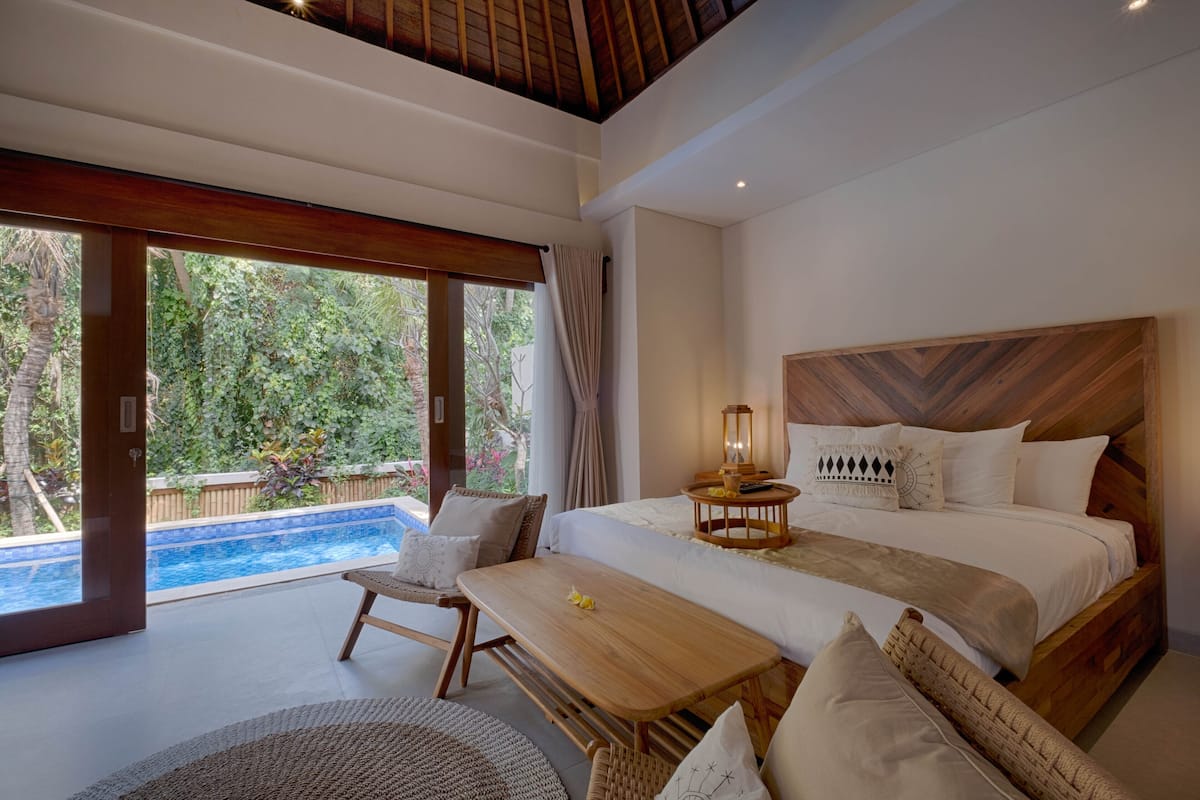 Quarto com cama grande, cabeceira de madeira, mesa de cabeceira com um candeeiro e vista para uma piscina.