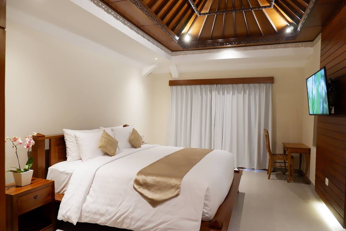 Quarto de hotel com uma cama grande, mesa de cabeceira de madeira, televisão na parede e uma janela com cortinas.
