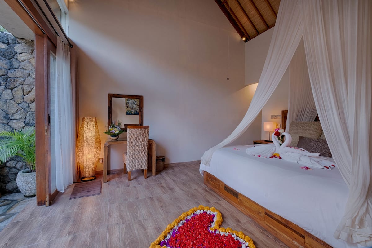 Quarto com cama de dossel, mesa de cabeceira com abajur, cadeira e um detalhe decorativo na parede.