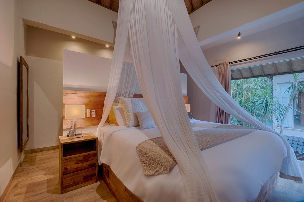 Quarto com cama de dossel, cabeceira de madeira, mesa de cabeceira com abajur e vista para o verde lá fora.