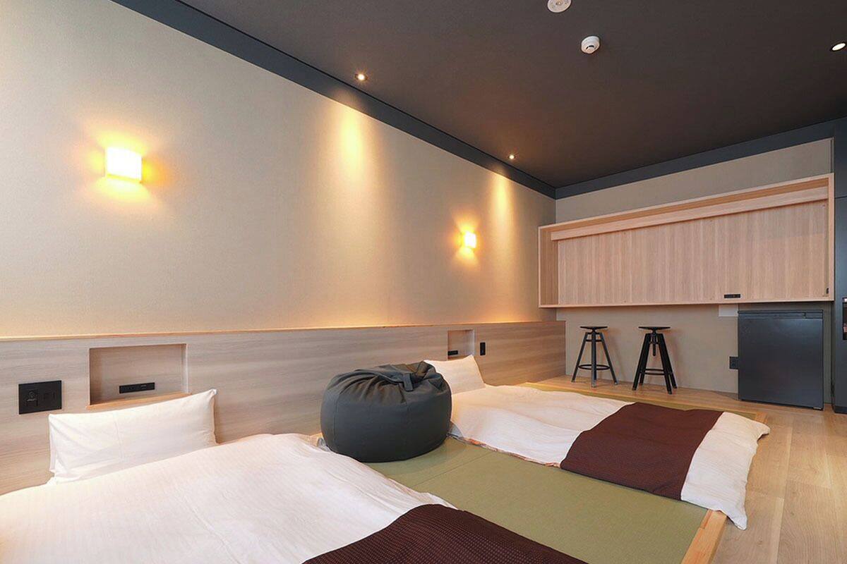 A modern hotel room with two beds, a bean bag, a kitchenette, and a large window.