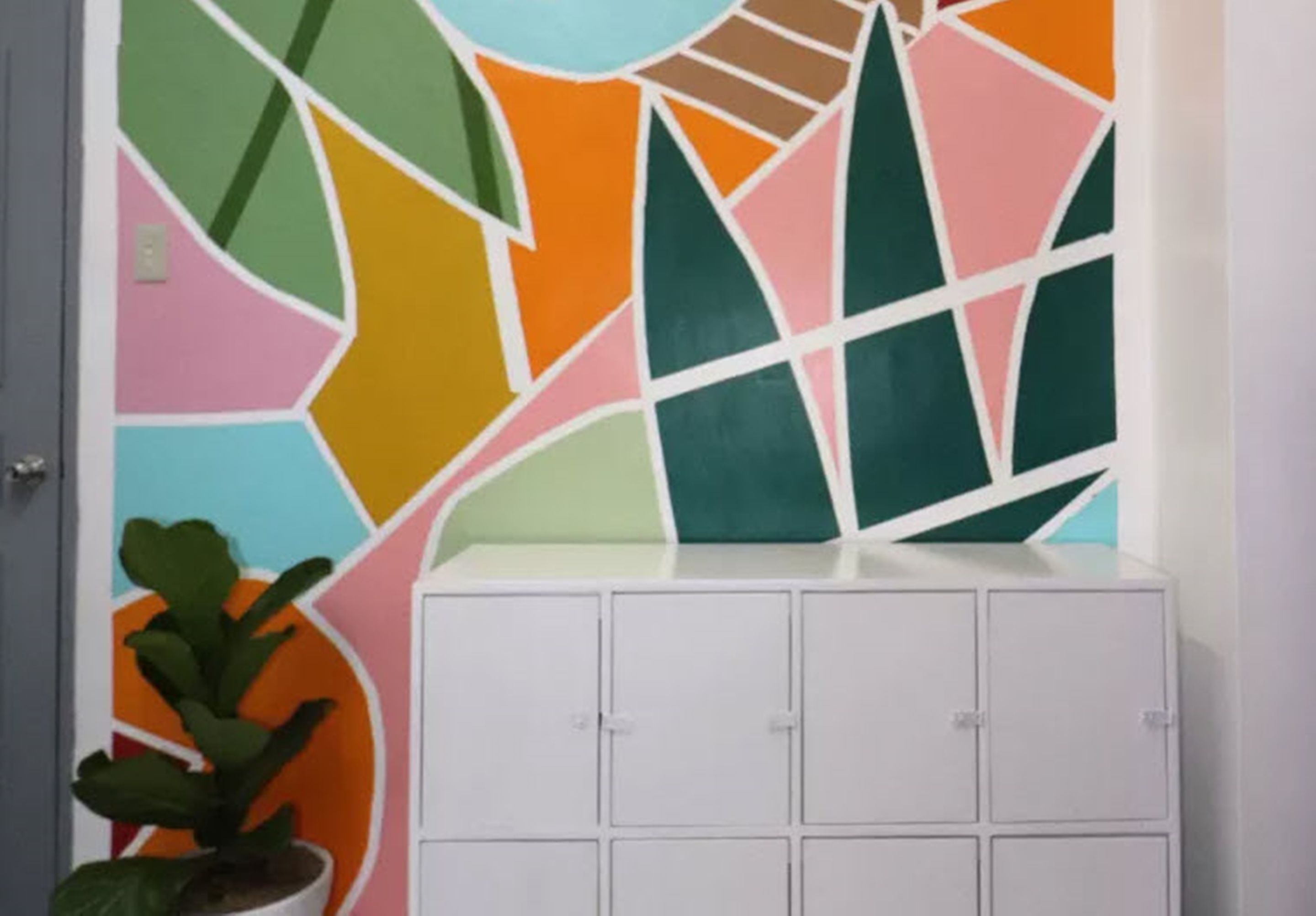 A room with a colorful abstract mural on the wall, a white cabinet, and a potted plant.
