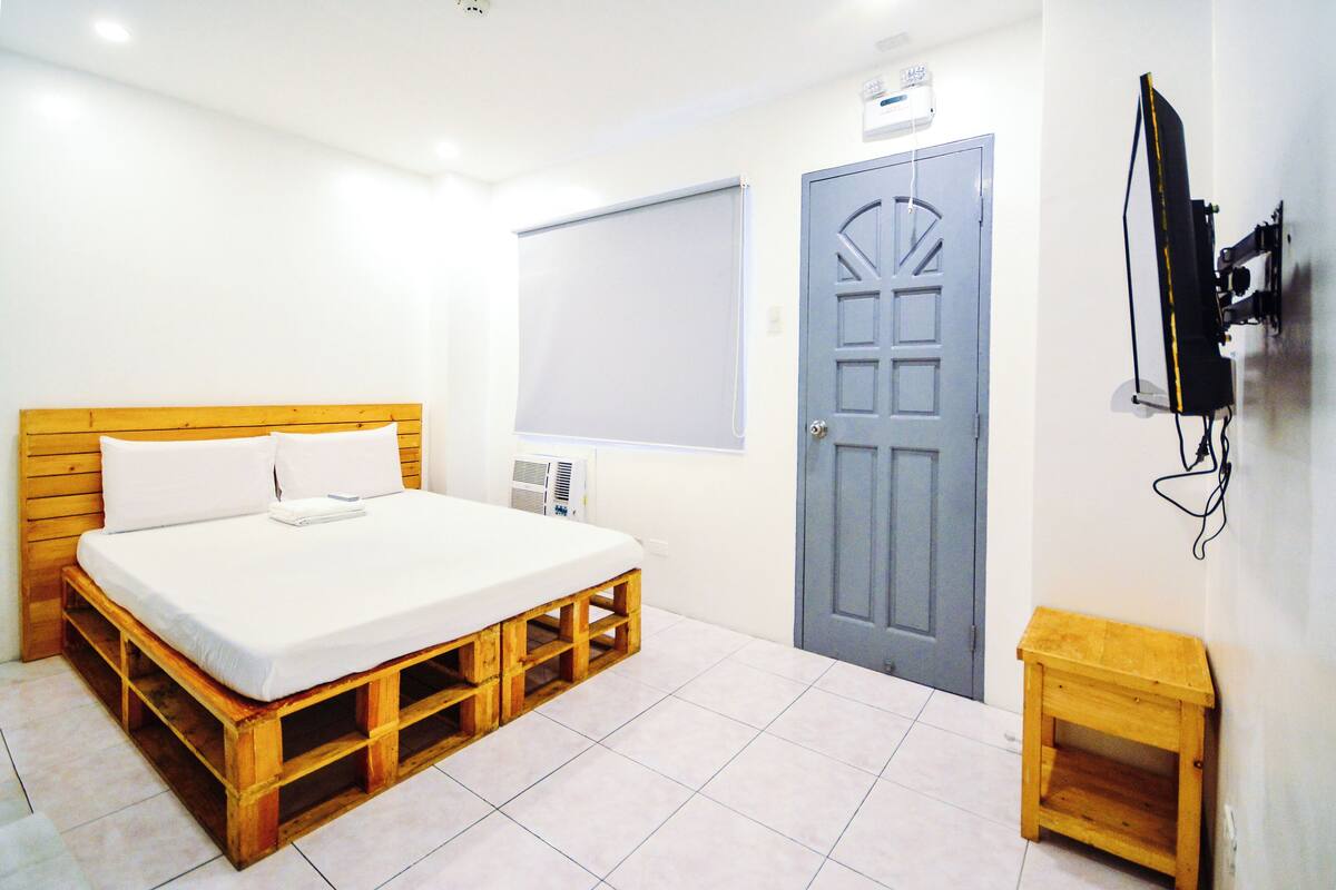 A bedroom with a wooden bed, a grey door, a white wall, and a mounted TV.
