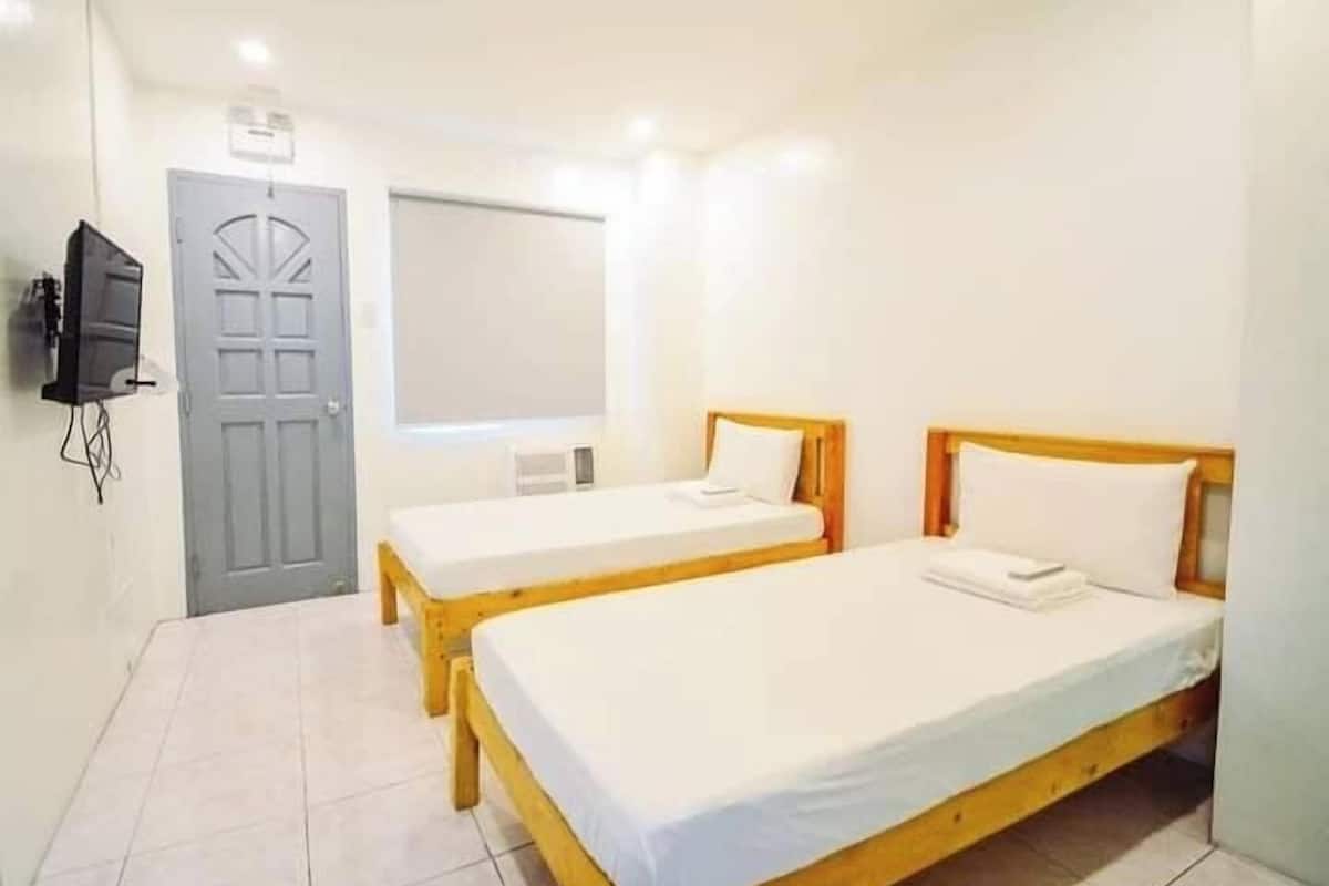 A room with two single beds, a grey door, a television mounted on the wall, and a window.