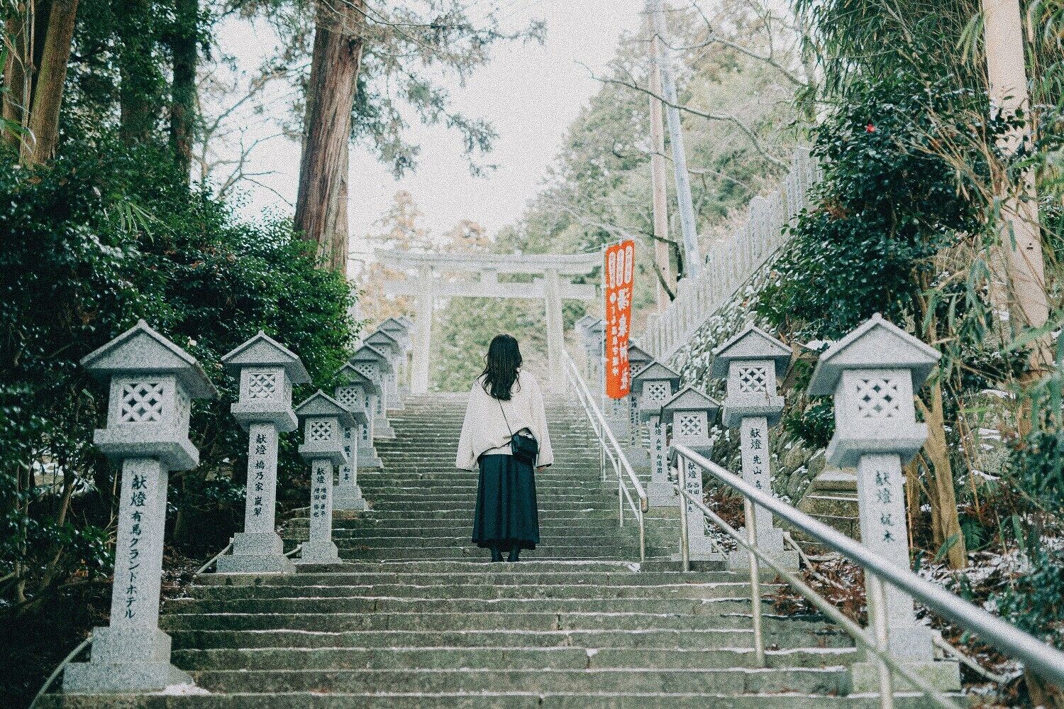 Una persona subiendo un tramo de escalones de piedra flanqueados por faroles tradicionales, en un entorno frondoso.