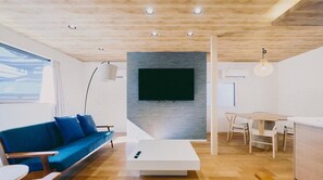 A modern living room with a blue sofa, a white coffee table, a dining table with chairs, a TV mounted on a grey wall, and a wooden ceiling. - Musubi no koyado enn by Wellbe - Hostel (Kobe)