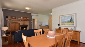 A living room with a fireplace, a dining table with chairs, a kitchen area, and a painting on the wall. - Somerville Wine Cottage Hideaway 3Bedroom Cottage (Broke)