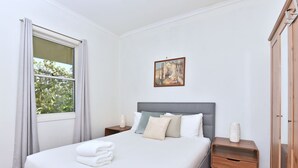 A neatly made bed with white linens and a stack of towels, a bedside table with a lamp, and a painting on the wall. - Somerville Wine Cottage Hideaway 3Bedroom Cottage (Broke)