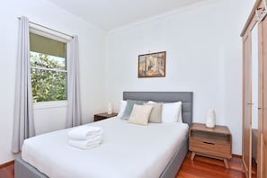 A neatly made bed with white linens and a stack of towels, a bedside table with a lamp, and a painting on the wall.