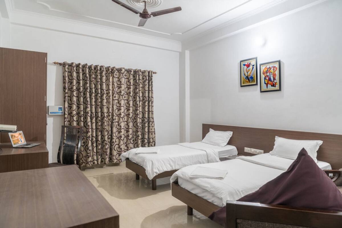 A hotel room with two single beds, a desk with a laptop, a ceiling fan, and two wall-mounted paintings.