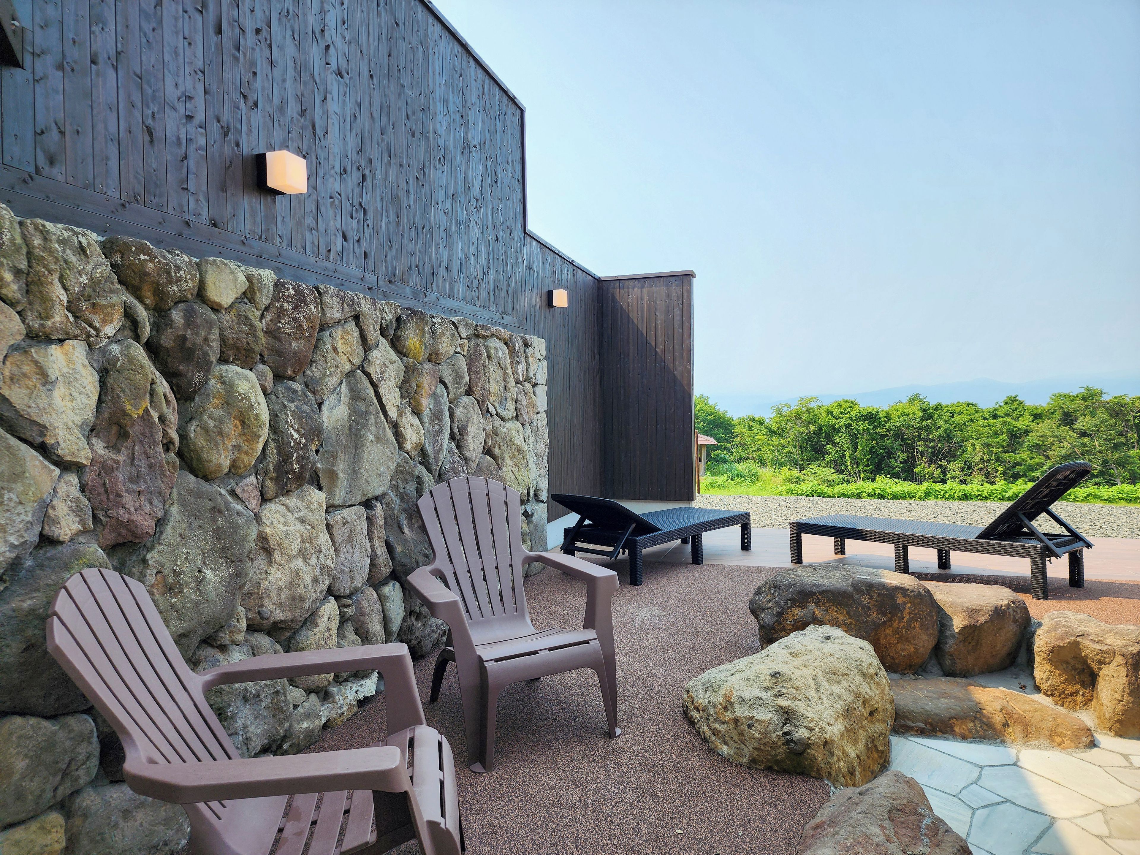 Un área de estar al aire libre con sillas Adirondack y un televisor de pantalla plana contra un muro de piedra, con vistas a un paisaje exuberante.