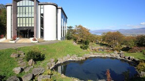 A modern building with a large glass facade, a stone pathway leading to the entrance, and a pond in the foreground. - Iwanai Kogen Hotel (Iwanai)