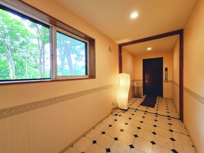 A hotel corridor with a tiled floor, a window with a view of trees, and a wall-mounted light fixture. - Iwanai Kogen Hotel (Iwanai)