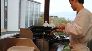 A person cooking in a kitchen with a view of a building and mountains in the background. - Iwanai Kogen Hotel (Iwanai)