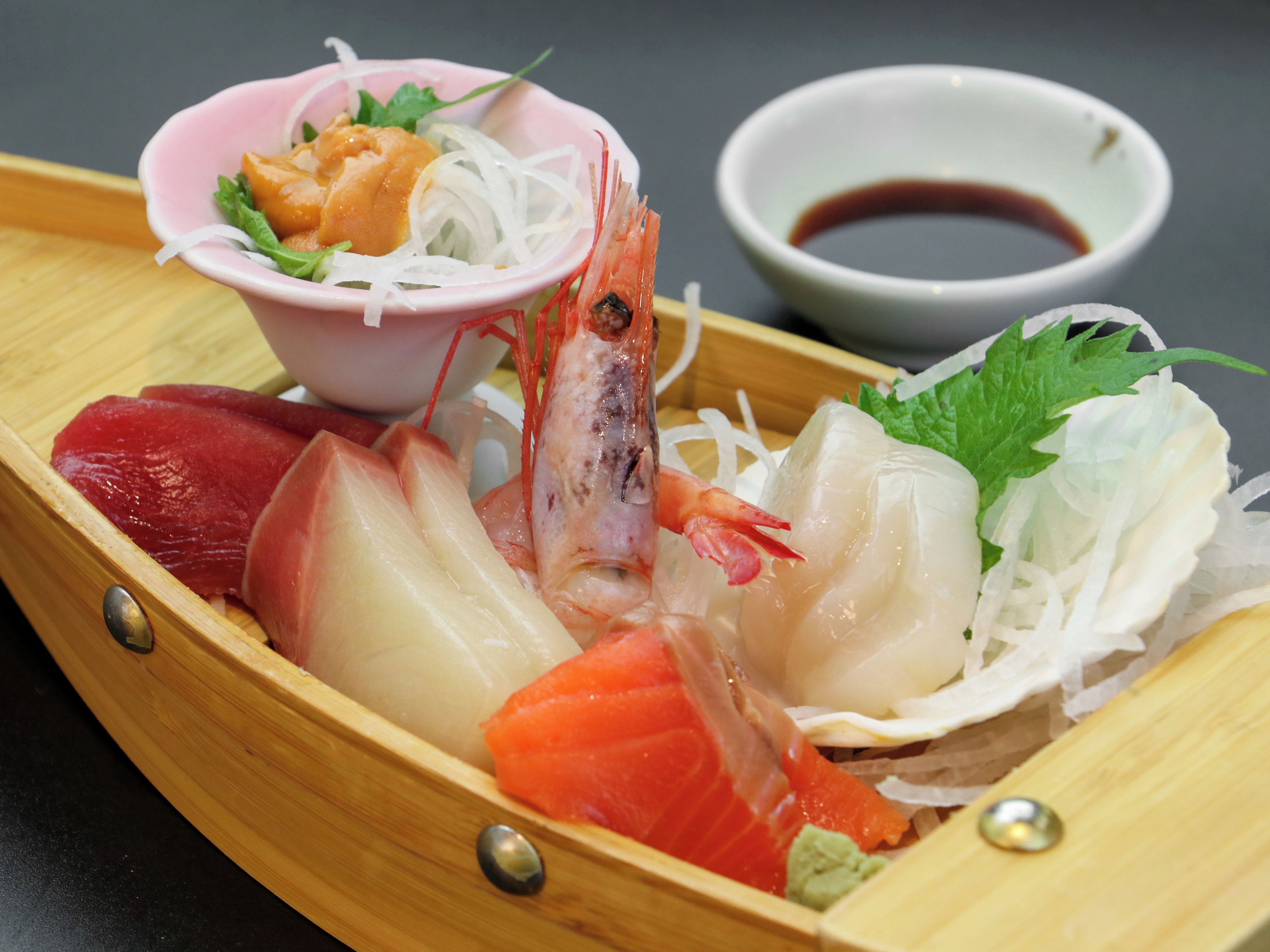 Een bootvormig houten serveerschaal met verschillende sashimi en een klein schaaltje sojasaus.