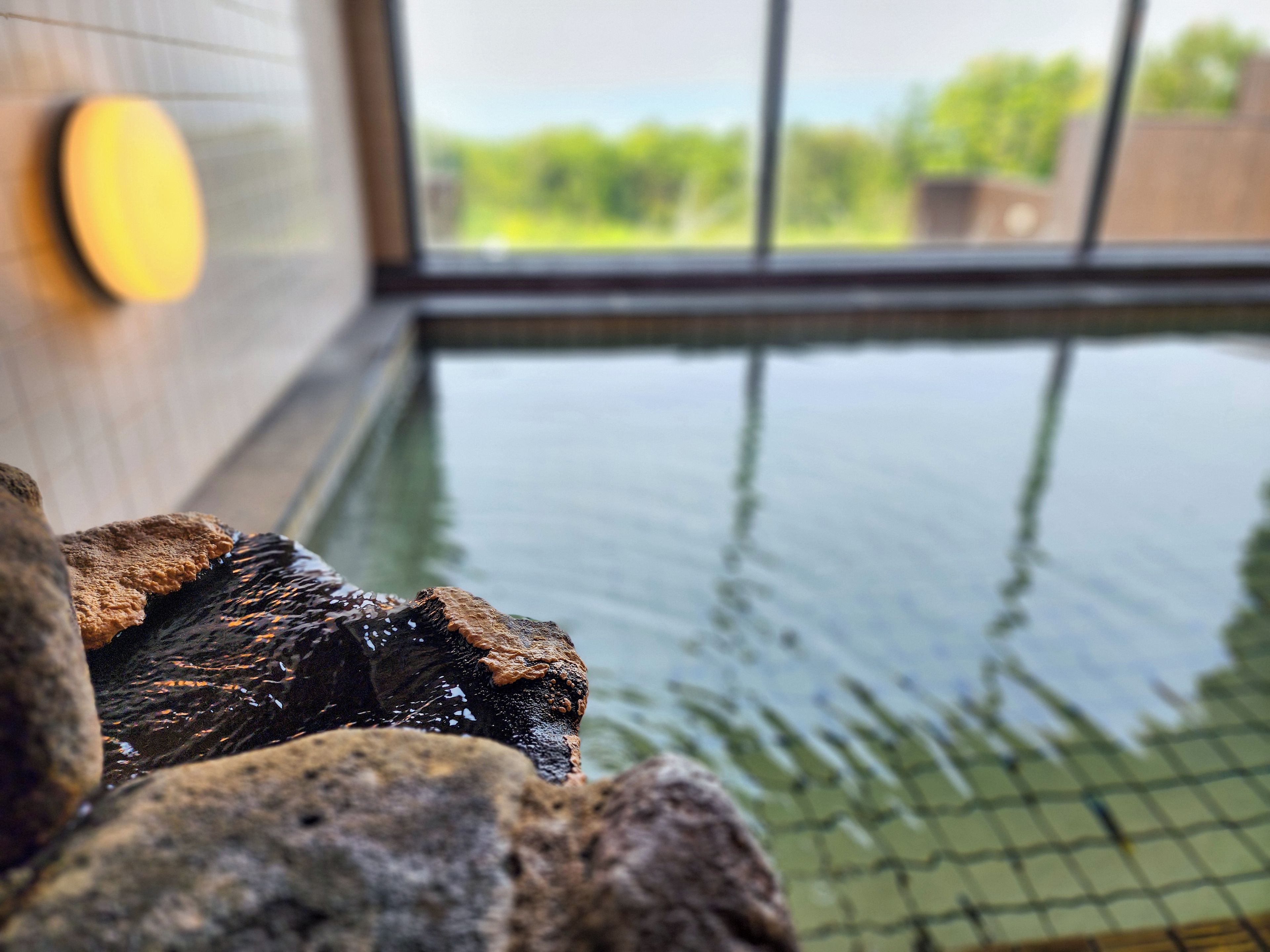 A stone wall with a water feature, a window with a view of greenery, and a tiled floor.