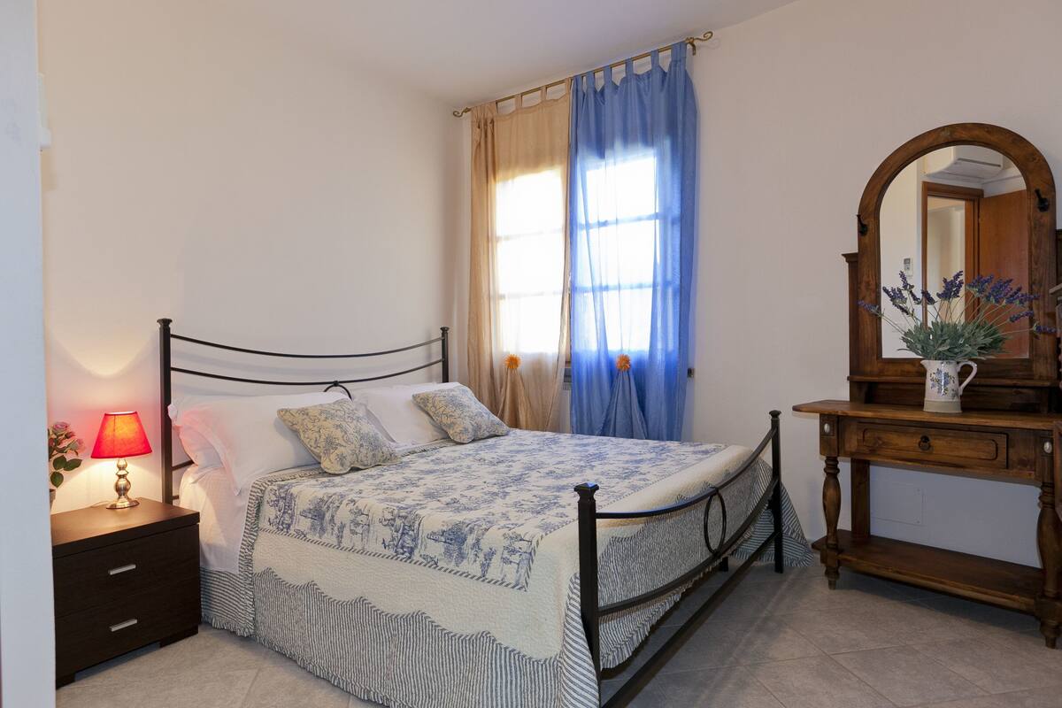 A bedroom with a bed, nightstand, lamp, dresser, mirror, and window with curtains.