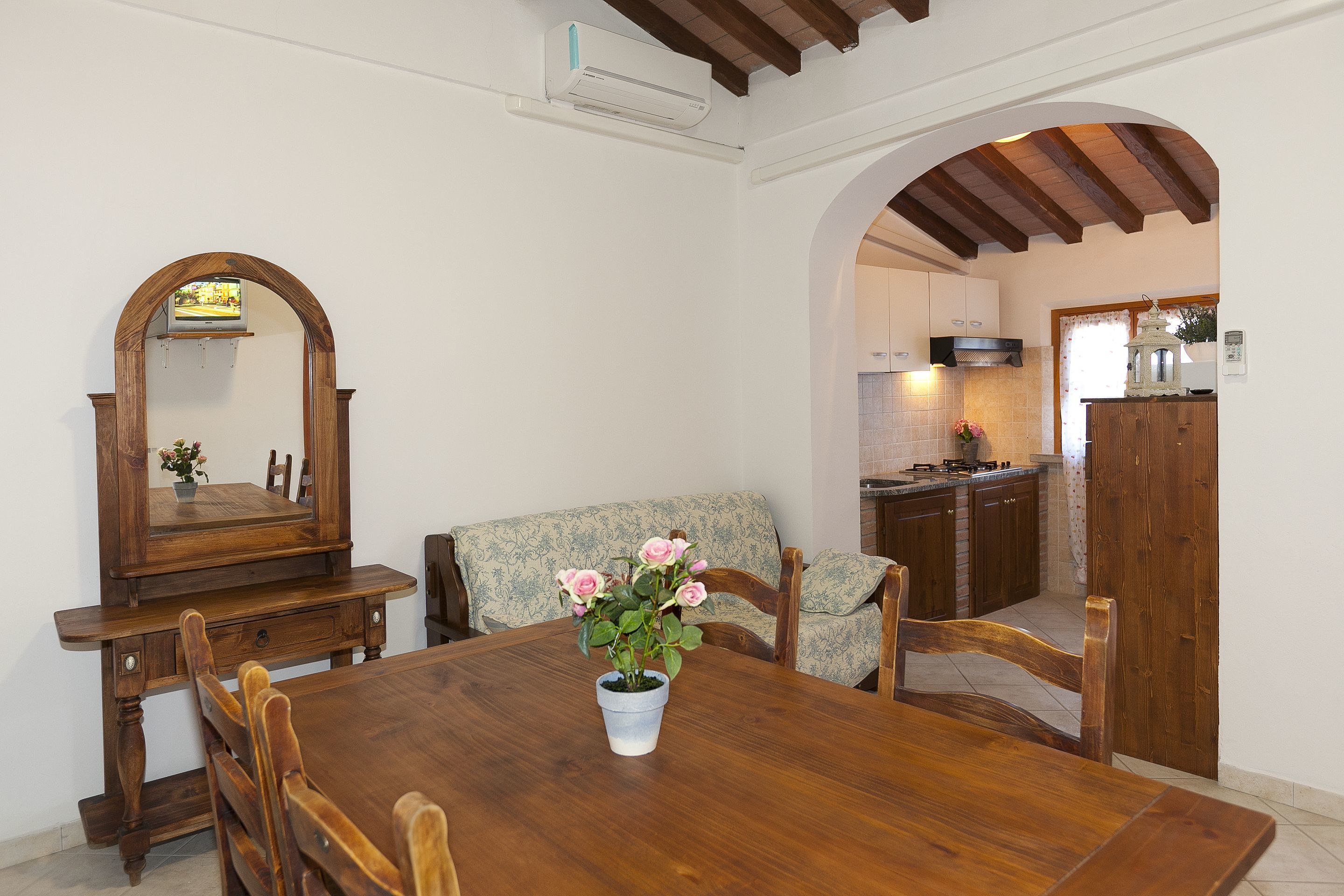 A dining area with a wooden table, chairs, a vase of flowers, a wooden bench, and a kitchen in the background.