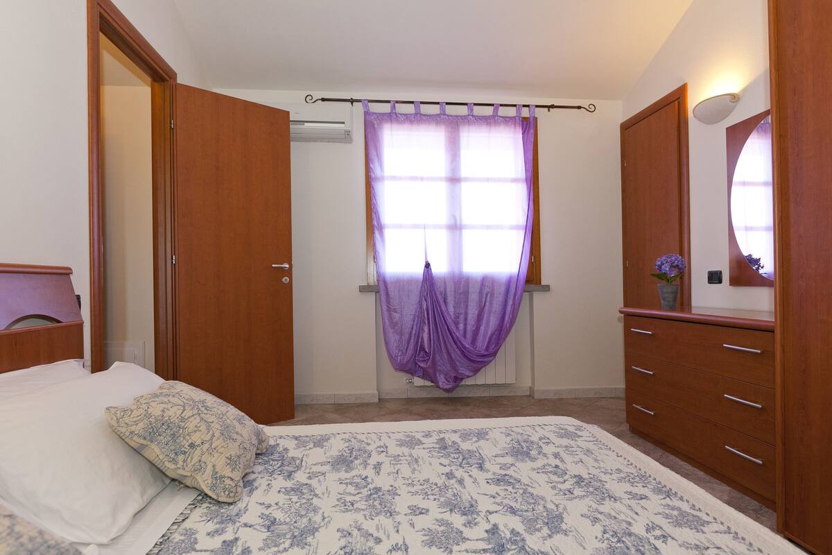 A bedroom with a bed, a dresser, a mirror, and a window with purple curtains.
