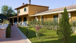 Ein zweistöckiges Gebäude mit überdachter Terrasse, einem kleinen Garten mit blühenden Pflanzen und einem gepflasterten Weg.
