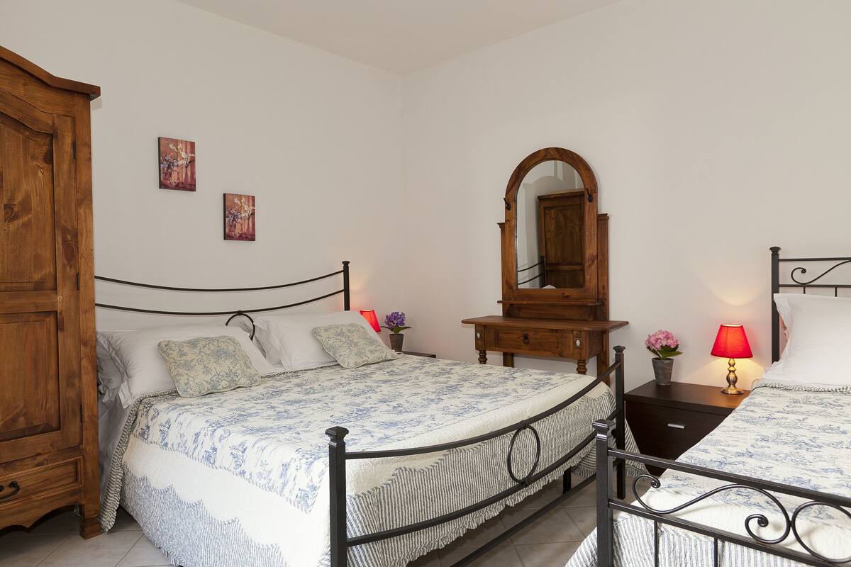 A bedroom with a bed, a wooden wardrobe, a nightstand with a lamp, and a mirror on the wall.