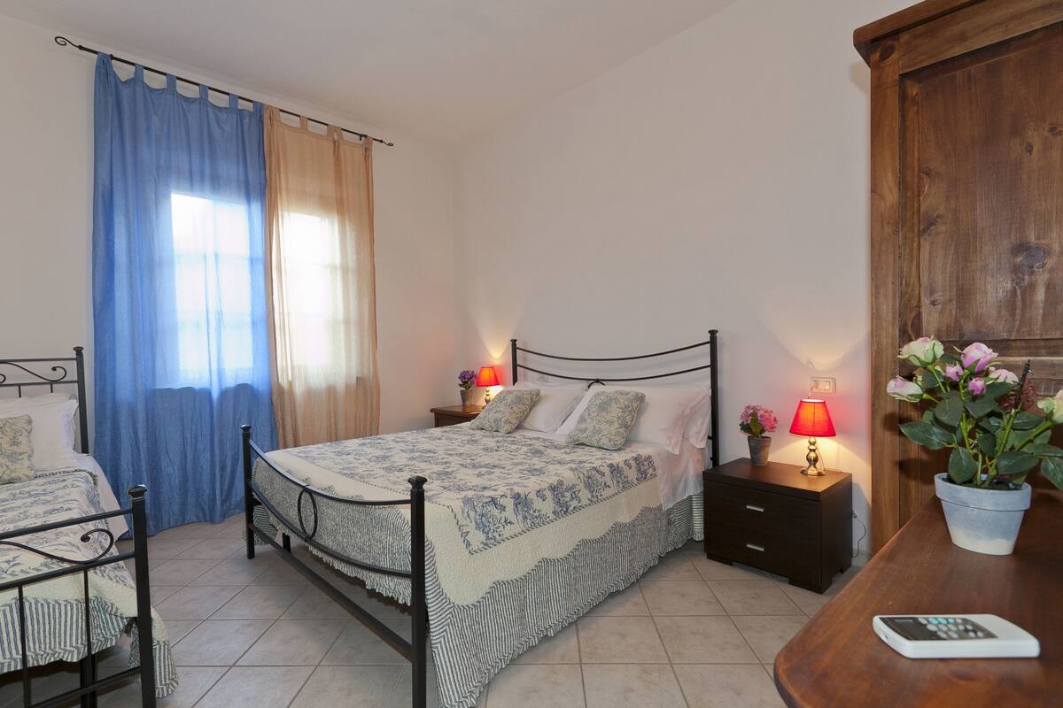 A bedroom with a bed, two bedside tables, a nightstand, a chair, a wooden cabinet, and a potted plant.