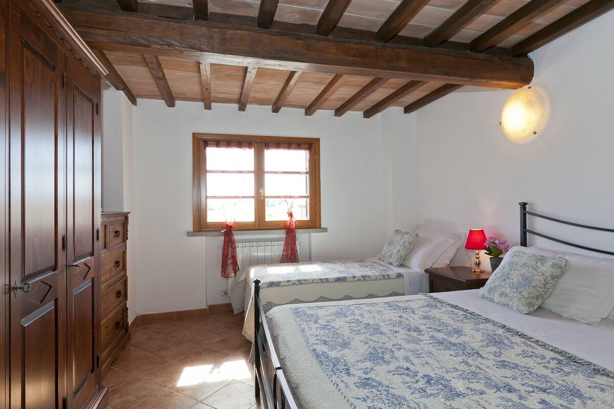 A bedroom with two beds, wooden wardrobe, a window with curtains, and a nightstand with a lamp.