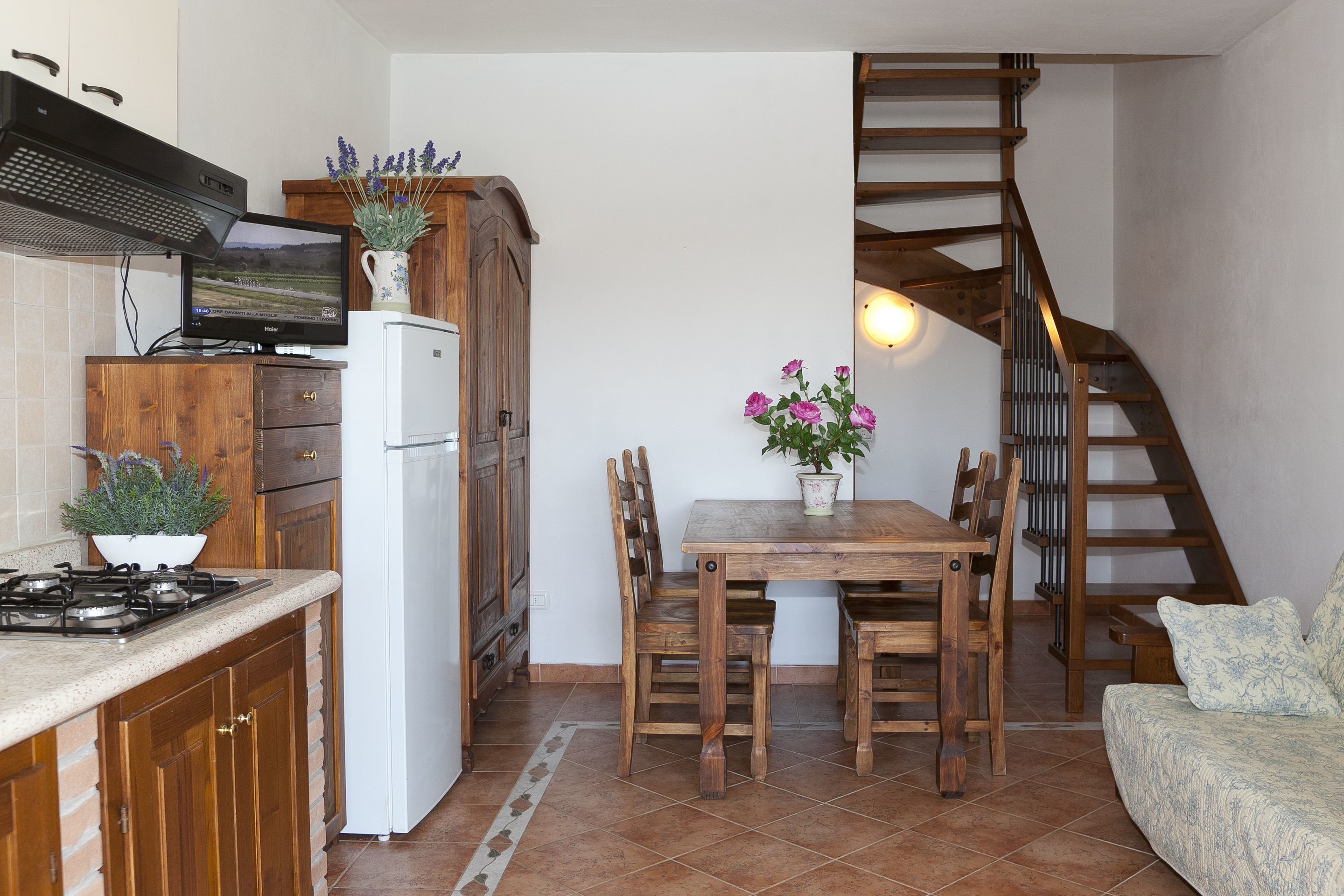 A compact kitchen with wooden cabinets, a white refrigerator, a stove, and a dining table with chairs. A staircase with wooden railings is visible in the background.