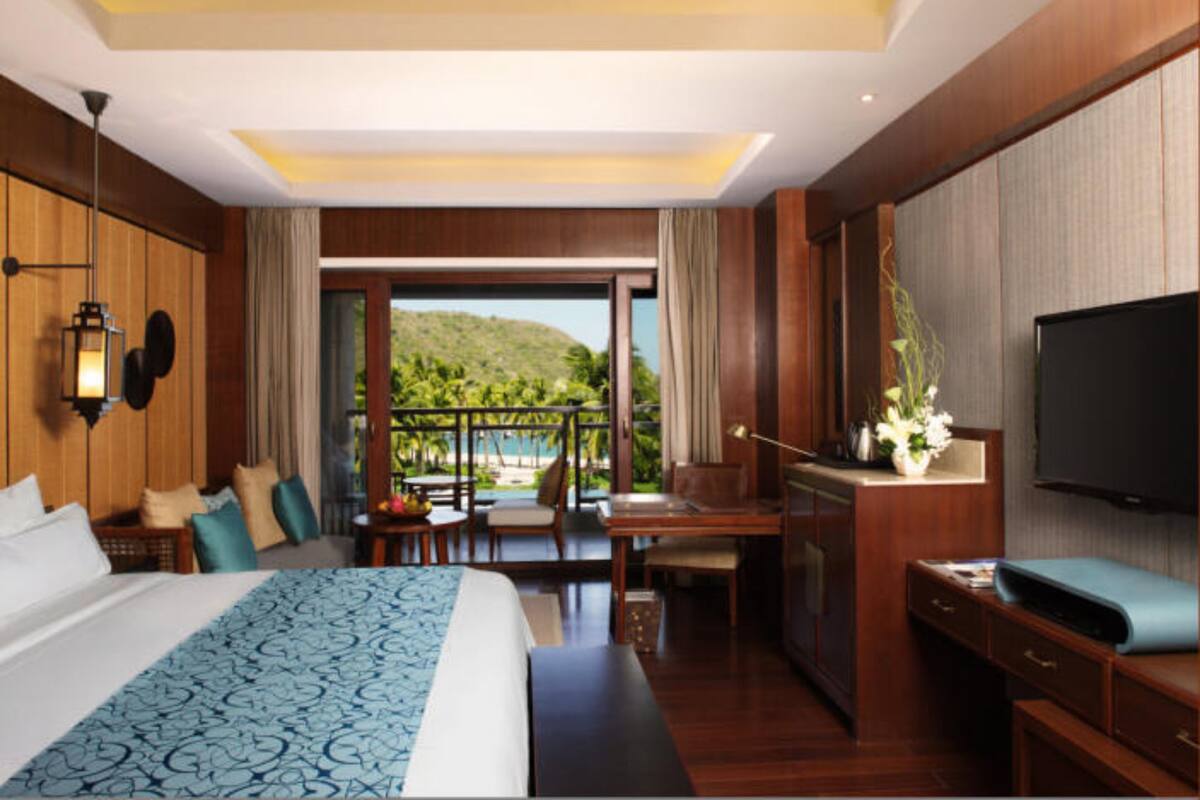 A hotel room with a large bed, a desk, a TV, and a balcony with a view of the hills.