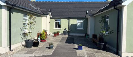 A courtyard with potted plants and a pathway leading to a house with a dark roof and light walls.