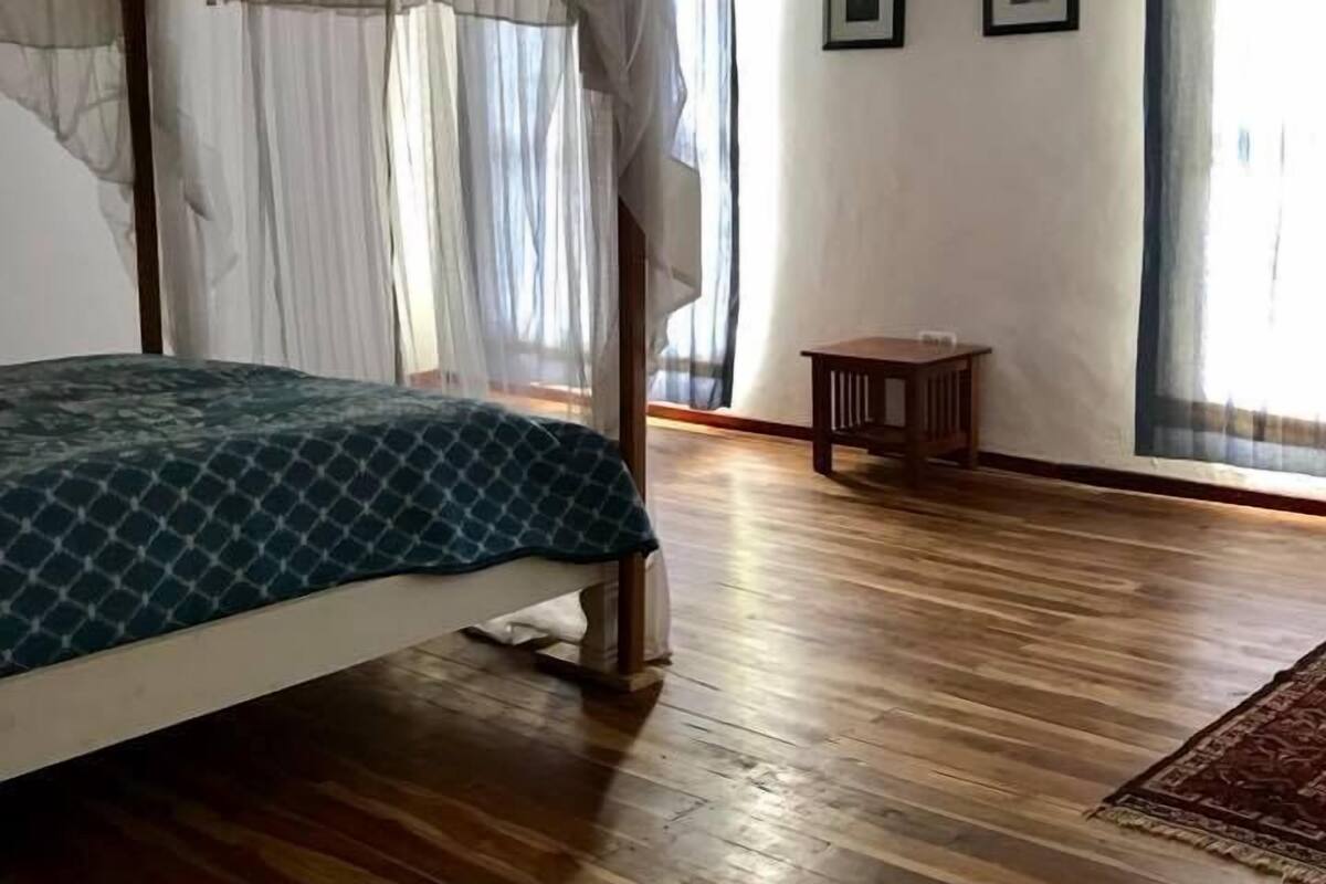 A bedroom with a four-poster bed, wooden flooring, a small table, and two framed pictures on the wall.