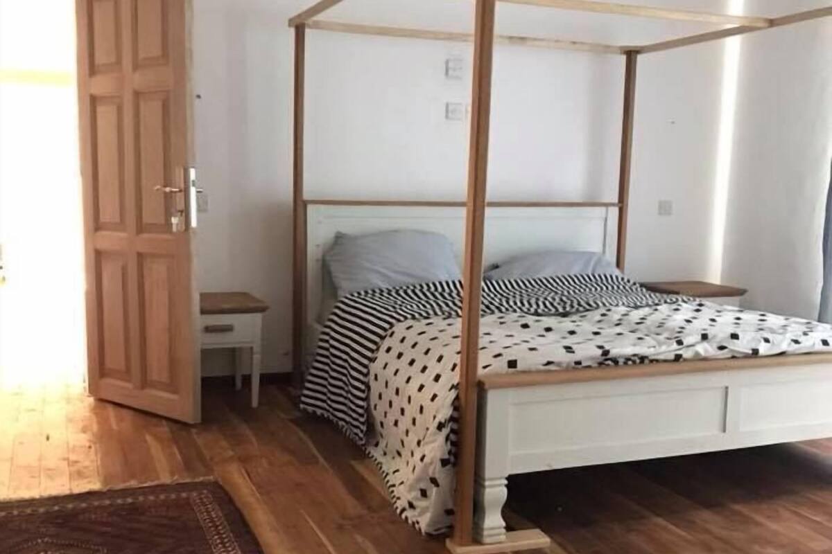 A room with a four-poster bed, a small bedside table, a patterned rug, and a wooden door.