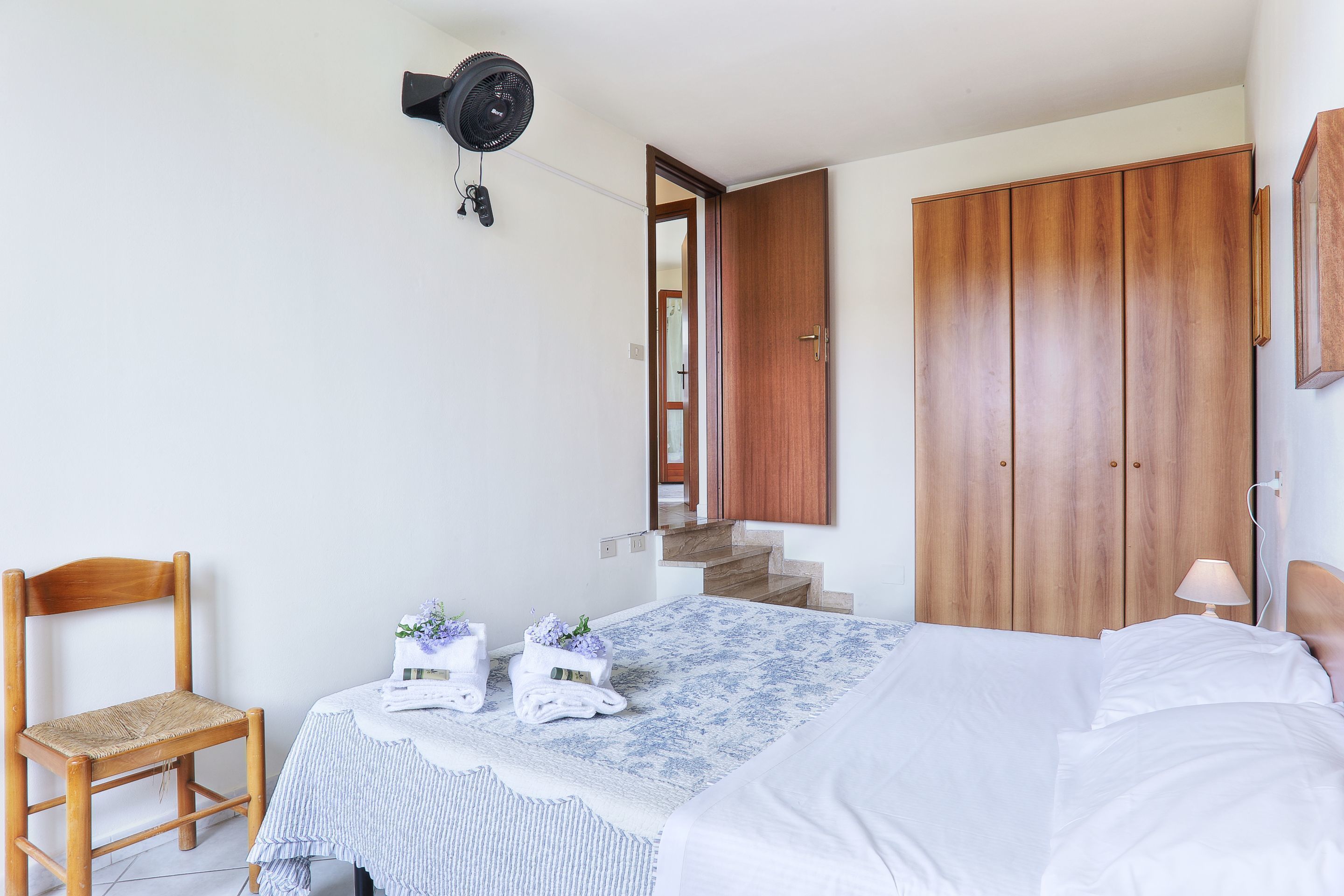 A bedroom with a bed, a chair, a wooden wardrobe, a staircase, and a wall-mounted fan.