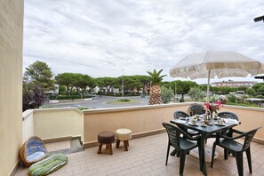 A balcony with a table, chairs, and an umbrella. There are two stools and a wicker chair. The balcony overlooks a street with trees and buildings.
