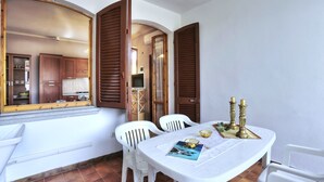 A dining area with a white table, two white chairs, and a view of a kitchen through a window with wooden shutters. - Case Sobrini (Rosignano Marittimo)
