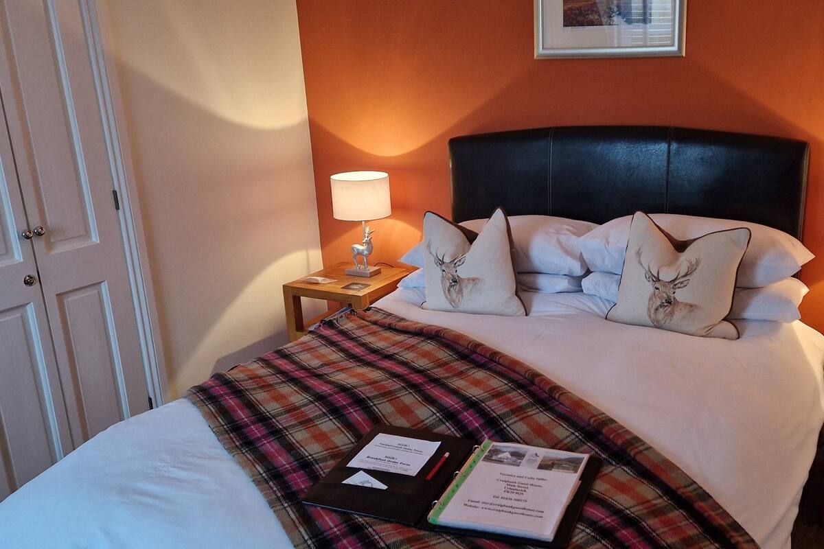 A hotel room with a bed, two bedside tables, a nightstand, a plaid blanket, and a book on the bed.