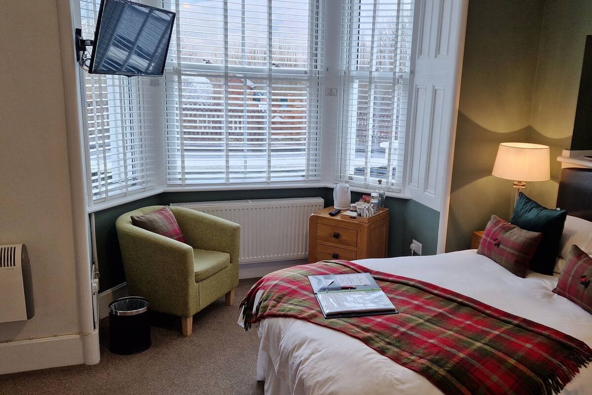 A bedroom with a bed, a chair, a TV, a lamp, and a window with blinds.