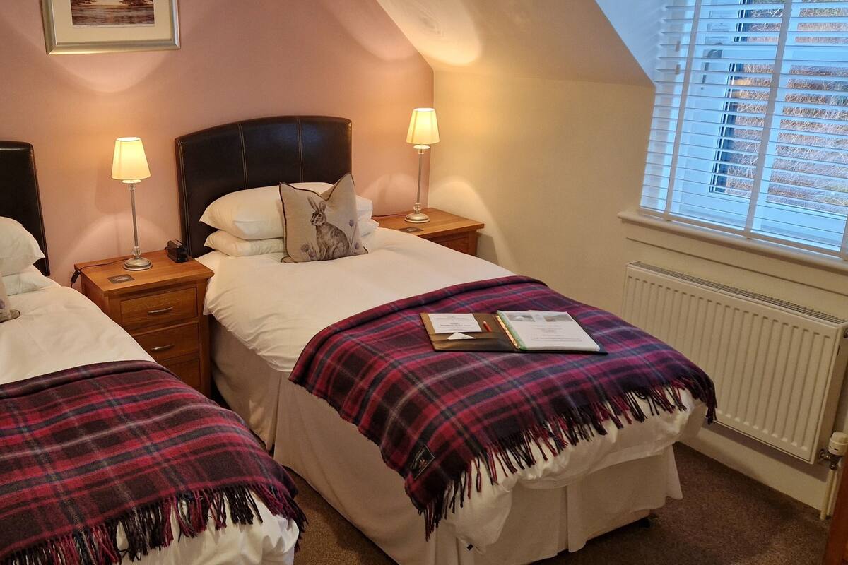 A cozy bedroom with two beds, each with a pillow featuring a rabbit design, a bedside table with a lamp, and a window with blinds.