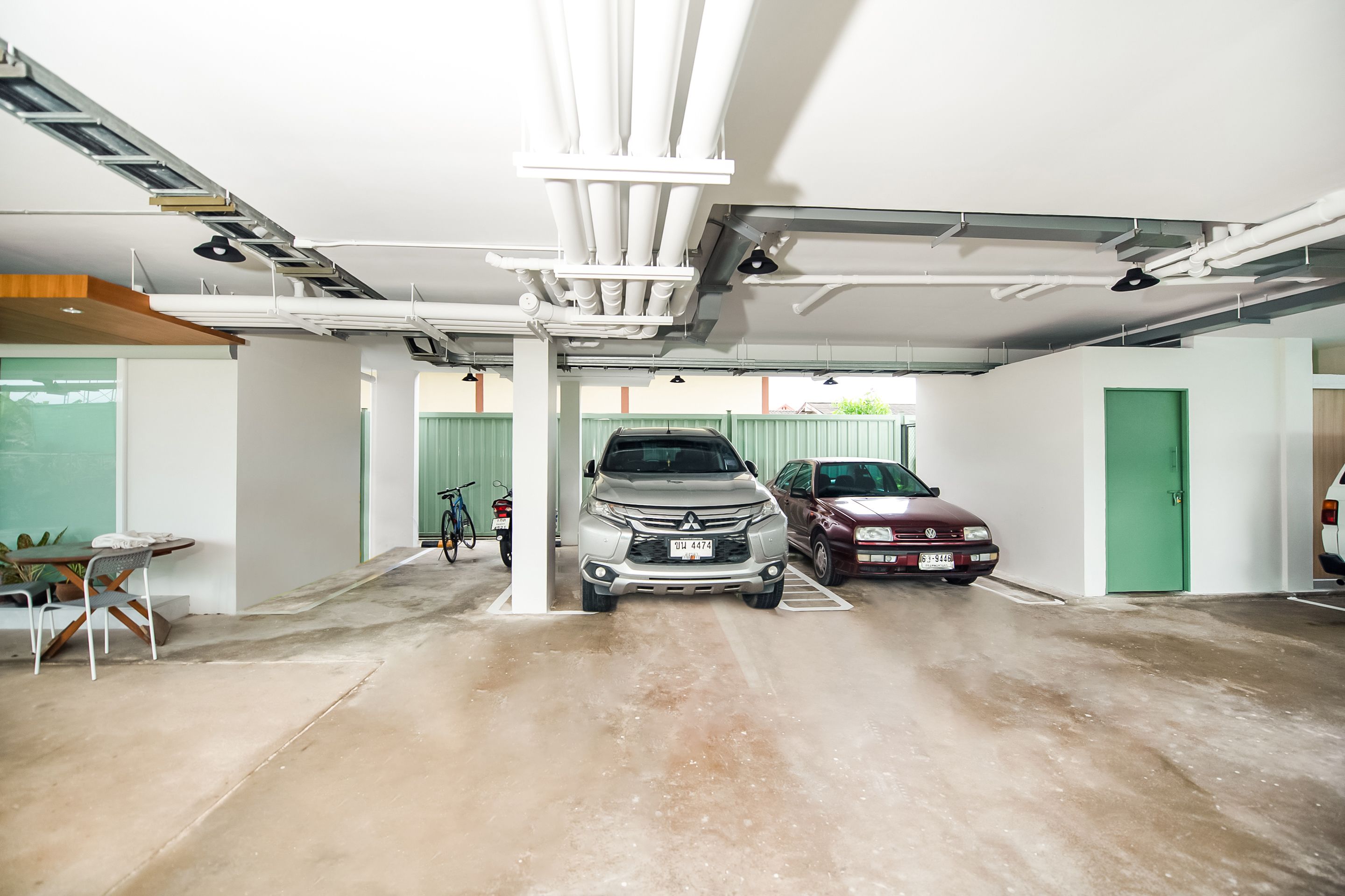 A parking garage with concrete floors and white walls, featuring two parked cars and a table with chairs.