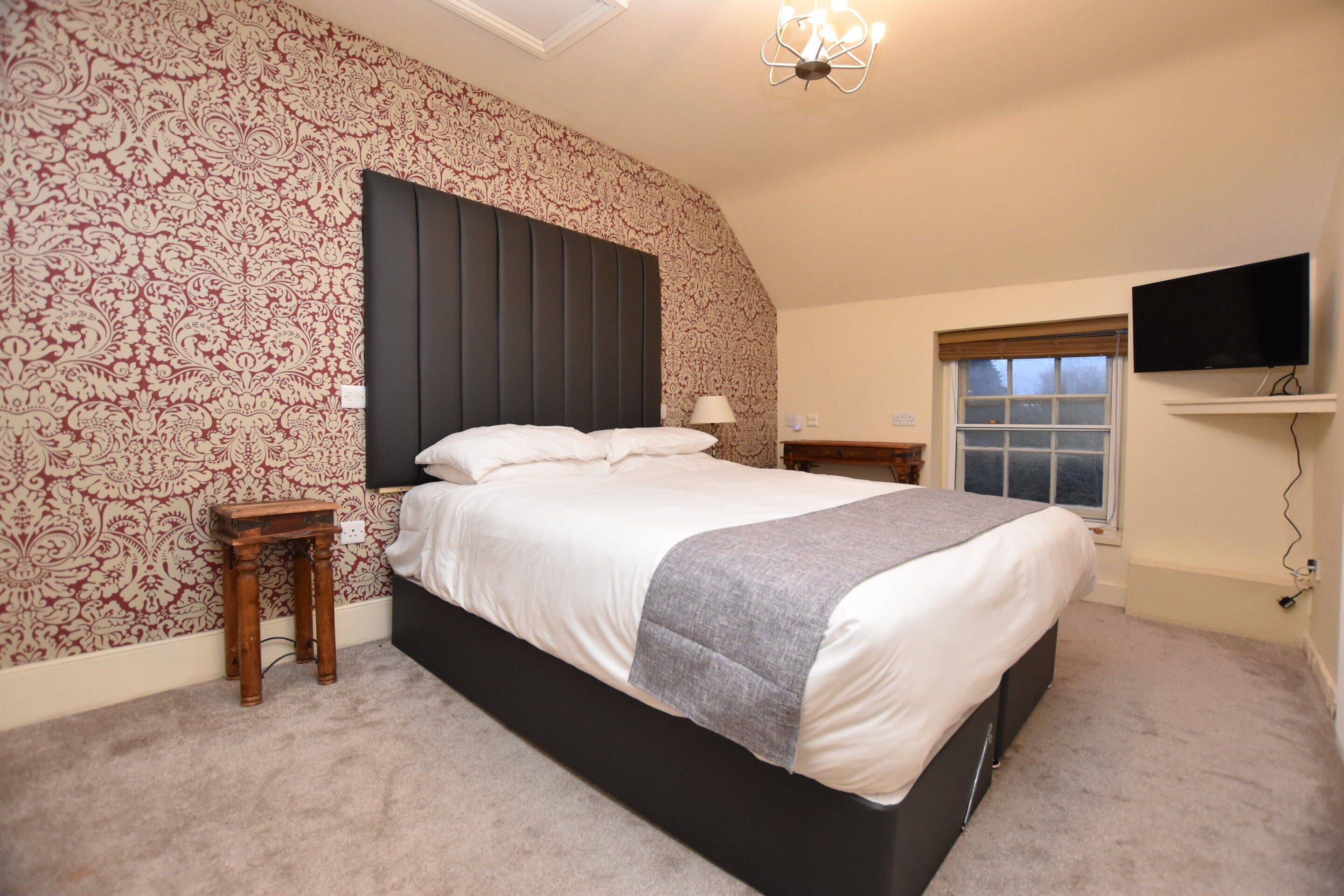 Une chambre avec un grand lit, une table de chevet en bois, une commode, une lampe, un téléviseur fixé au mur et une fenêtre avec des stores.