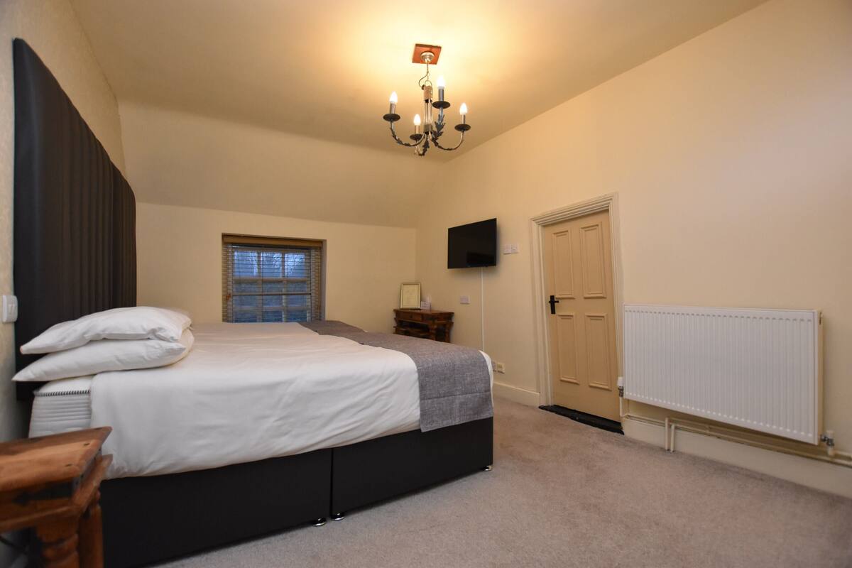 A bedroom with a bed, a wooden nightstand, a TV mounted on the wall, a window with blinds, and a door.