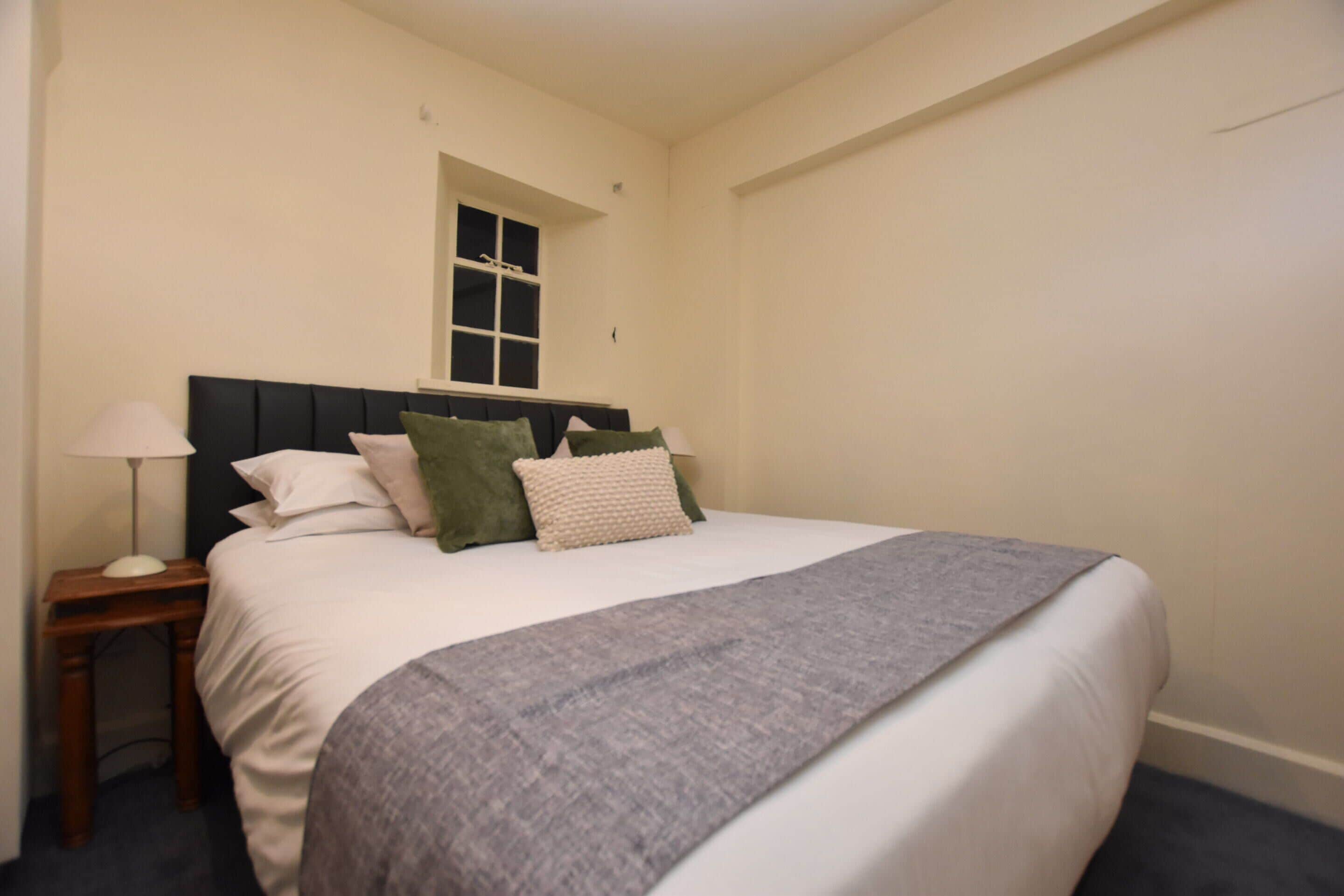 Een net opgemaakt bed met een grijze deken, een nachtkastje met een lamp en een raam boven het bed.