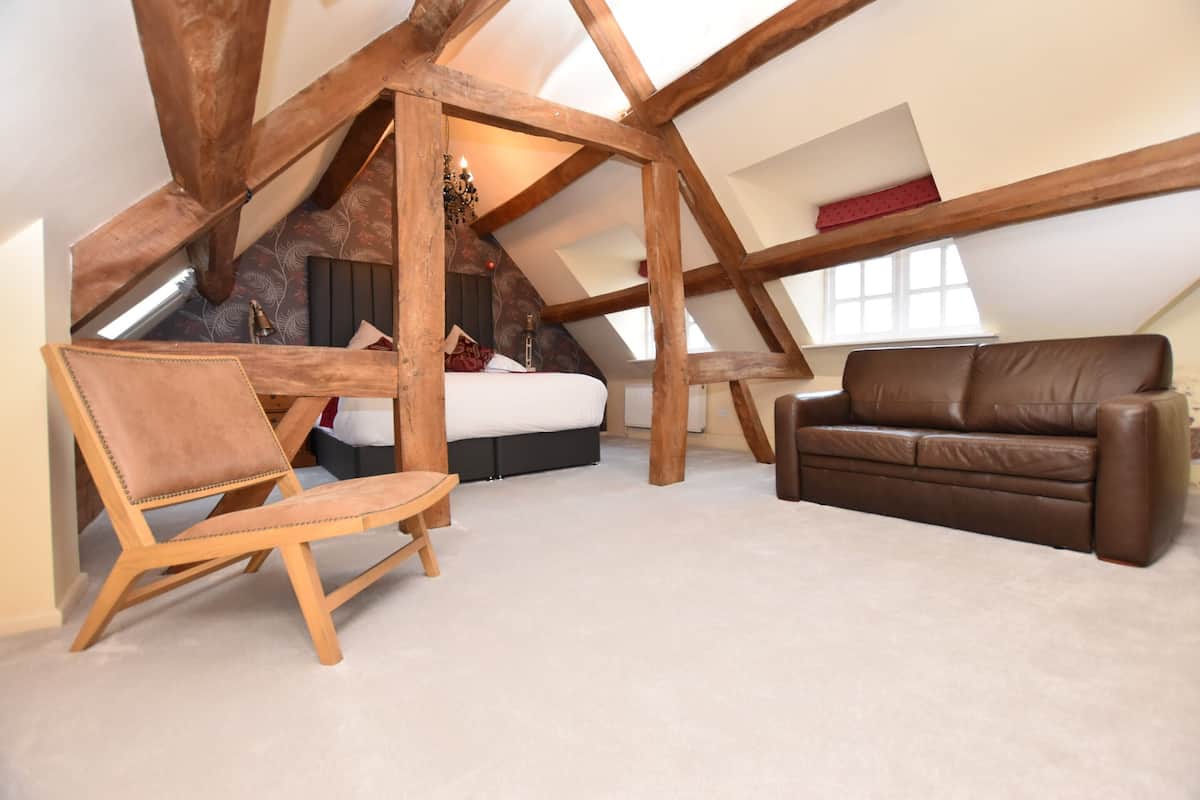 A loft bedroom with a sloped ceiling, wooden beams, a bed, a brown leather sofa, and a wooden chair.